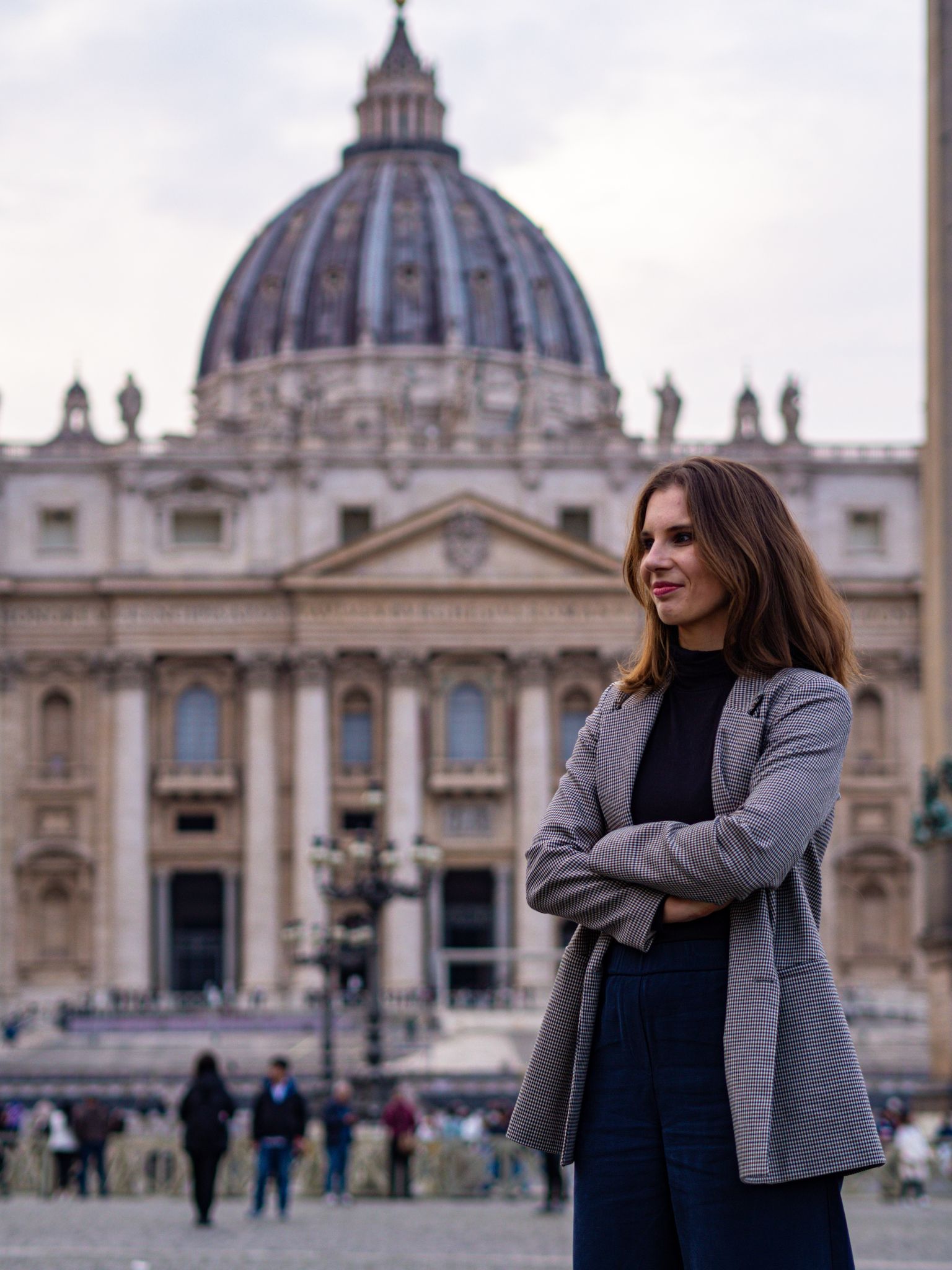Młoda kobieta w żakiecie ze skrzyżowanymi rękami stoi bokiem w Placu św. Piotra; w tle rozmyta fasada bazyliki i przechodnie